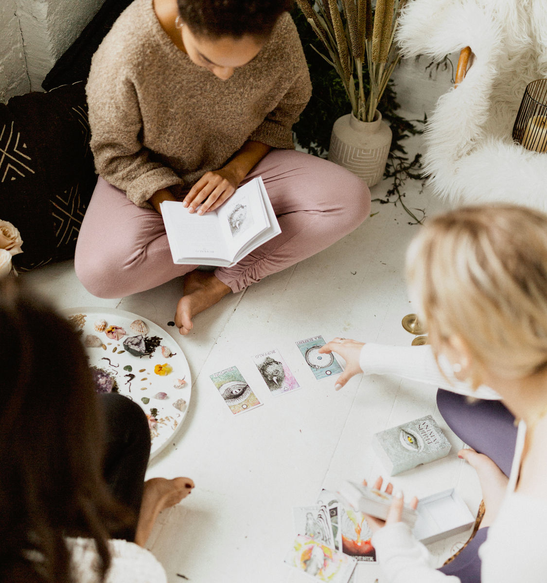 Three people in a cozy boho living room sitting cross-legged doing a tarot/oracle card reading with crystals and a booklet.
