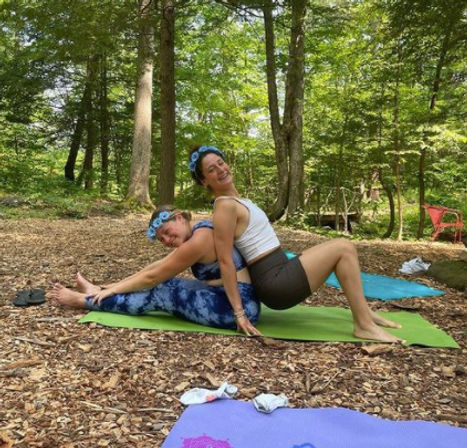 Two women doing partner yoga back-to-back on green mats in a sunlit forest clearing, wearing athletic clothes and floral headbands, surrounded by trees and leaf-strewn ground