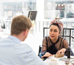 Woman with headscarf and tattoos reading tarot cards for a man at a sunlit urban café table, decks, crystals and a coffee cup visible