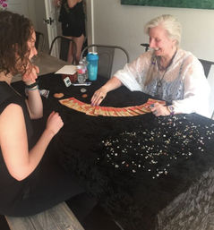 Smiling tarot reader lays a fanned spread of tarot cards across a black-clothed table scattered with crystals while a seated client looks on in a cozy indoor setting — tarot reading and crystal consultation.