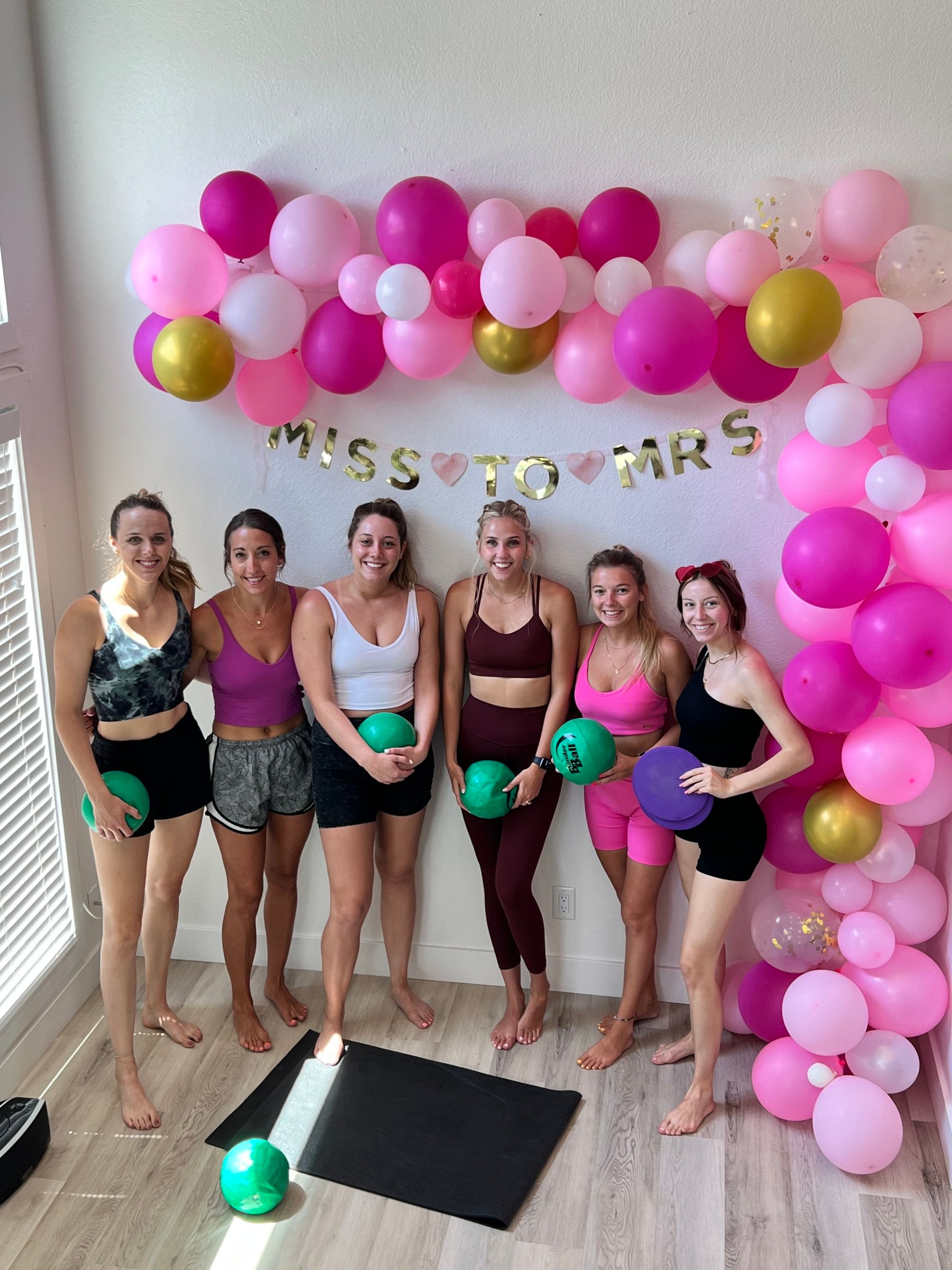 Six friends in colorful activewear pose barefoot in a bright room holding small fitness balls under a pink-and-gold balloon garland and “Miss to Mrs” banner — playful at-home bachelorette workout scene.