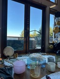 Sunlit cozy meditation setup by glass patio doors overlooking a deck and evergreen trees — crystal singing bowls, drum, cushions and circular stand on a wooden floor, with black and gold balloon garland at the window.