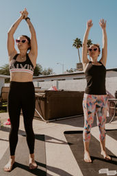 Two women practicing outdoor yoga on black mats in a sunny backyard patio with palm trees, wearing heart-shaped sunglasses—one in black leggings and a sports bra, the other in colorful patterned leggings—stretching arms overhead.