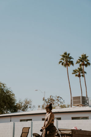 Woman in a sports bra and camouflage leggings standing in a sunny suburban backyard by lounge chairs and a low cinderblock wall, tall palm trees and clear blue sky overhead.