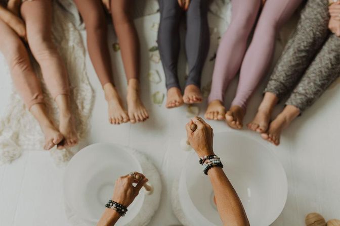 Top-down view of barefoot participants in colorful leggings seated in a circle in a bright wellness studio while a facilitator plays crystal singing bowls for a relaxing sound bath