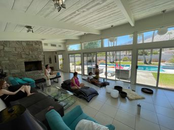 Sunlit mid-century living room with stone fireplace and floor-to-ceiling sliding glass doors opening to a backyard pool and palm trees; a small group relaxes on sofas and floor cushions.