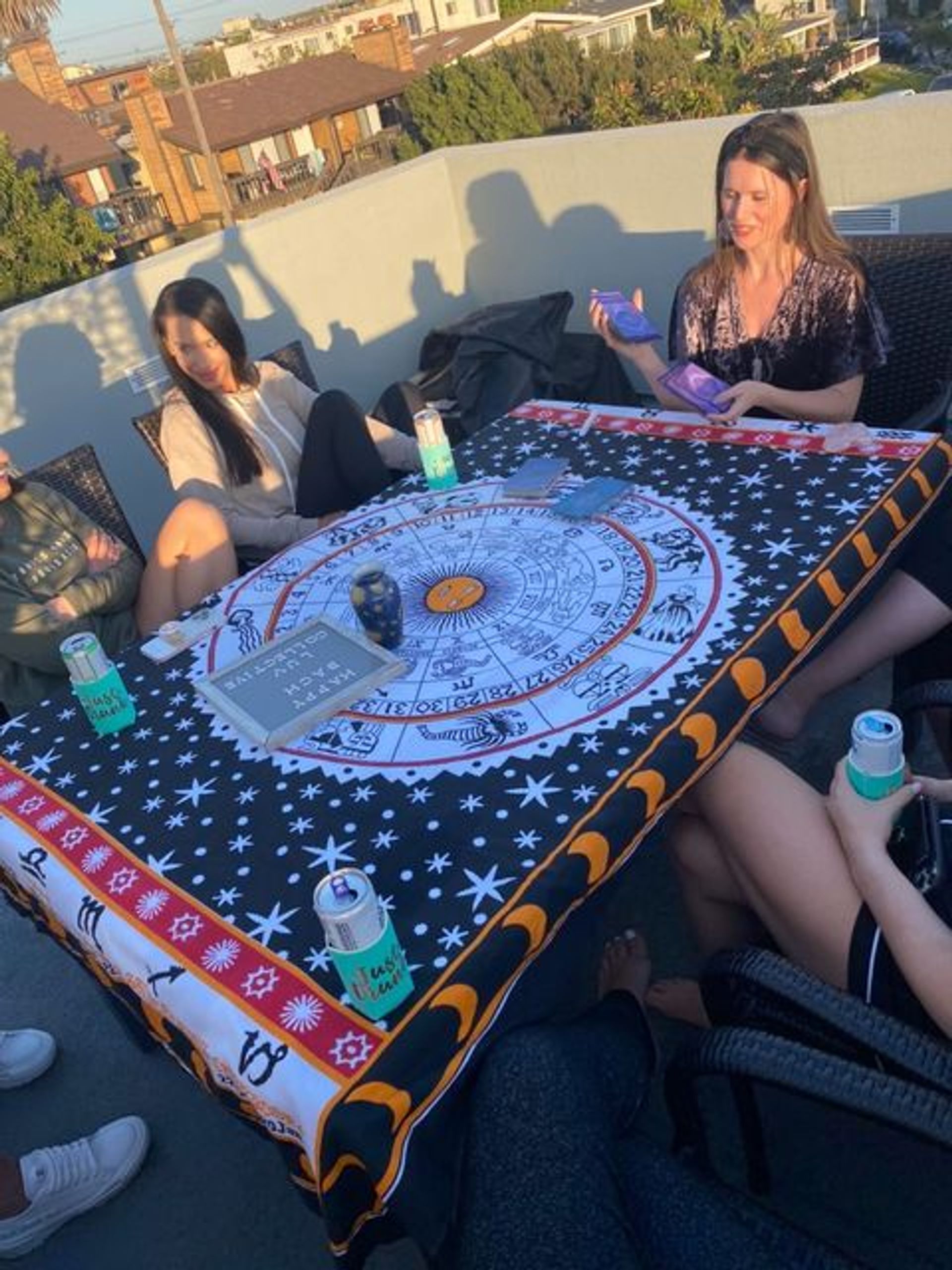 Small group on a rooftop deck at sunset gathered around a zodiac-themed tablecloth with tarot cards, candles and canned drinks for a casual astrology reading.