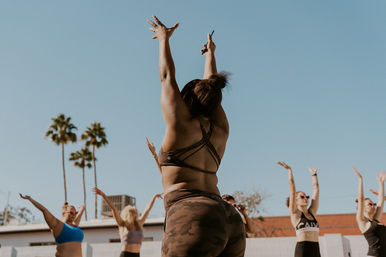 Energetic outdoor workout: instructor seen from behind in a strappy sports bra and camo leggings raises arms to a clear blue sky while participants mirror the pose, palm trees in the background.