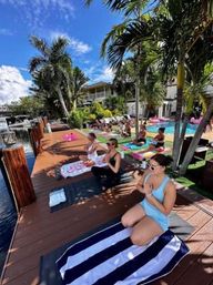 Sunny poolside yoga and meditation on a tropical waterfront dock, participants seated on mats and striped towels beside a pool and palm trees under a bright blue sky.