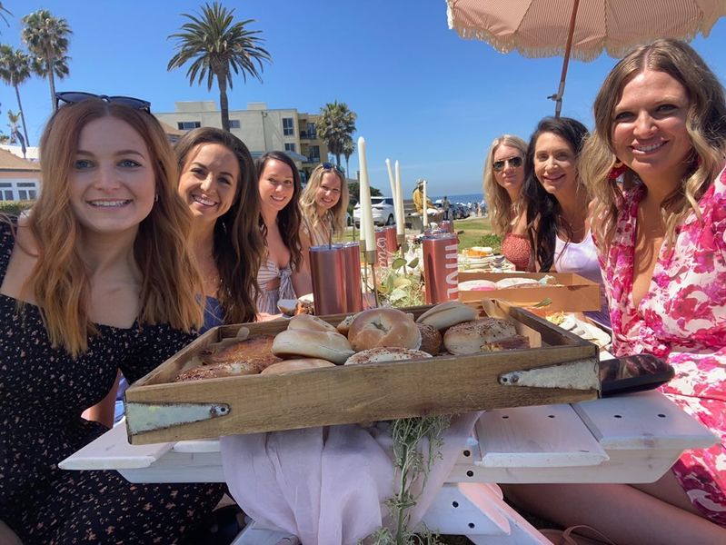 Seven women enjoying a sunny beachside brunch at a white picnic table, wooden tray of bagels in the foreground with palm trees and ocean view under a blue sky.