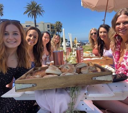 Seven women enjoying a sunny beachside brunch at a white picnic table, wooden tray of bagels in the foreground with palm trees and ocean view under a blue sky.