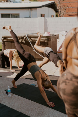 Three women balancing in three‑legged downward‑dog on black yoga mats in a sunny suburban backyard beside a hot tub — outdoor group yoga/fitness session with water bottle and phone nearby.
