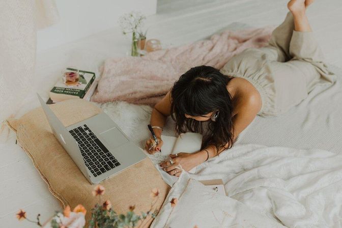 Person lying on a cozy bedroom workspace, laptop open on a pillow and writing in a notebook on soft blankets with floral accents