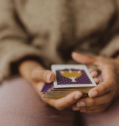 Close-up of hands holding a fanned tarot deck with the Three of Swords face-up, purple and gold card design and a cozy sweater background