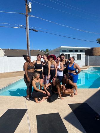Group of eleven women in activewear and heart-shaped sunglasses posing by a sunny suburban backyard pool with yoga mats on the deck