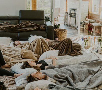 Sunlit modern living room with wood-paneled walls and fireplace; a group of people lying on pillows and blankets on the floor, relaxing in a cozy indoor retreat.
