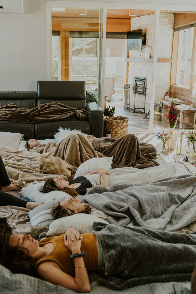 Sunlit modern living room with wood-paneled walls and fireplace; a group of people lying on pillows and blankets on the floor, relaxing in a cozy indoor retreat.