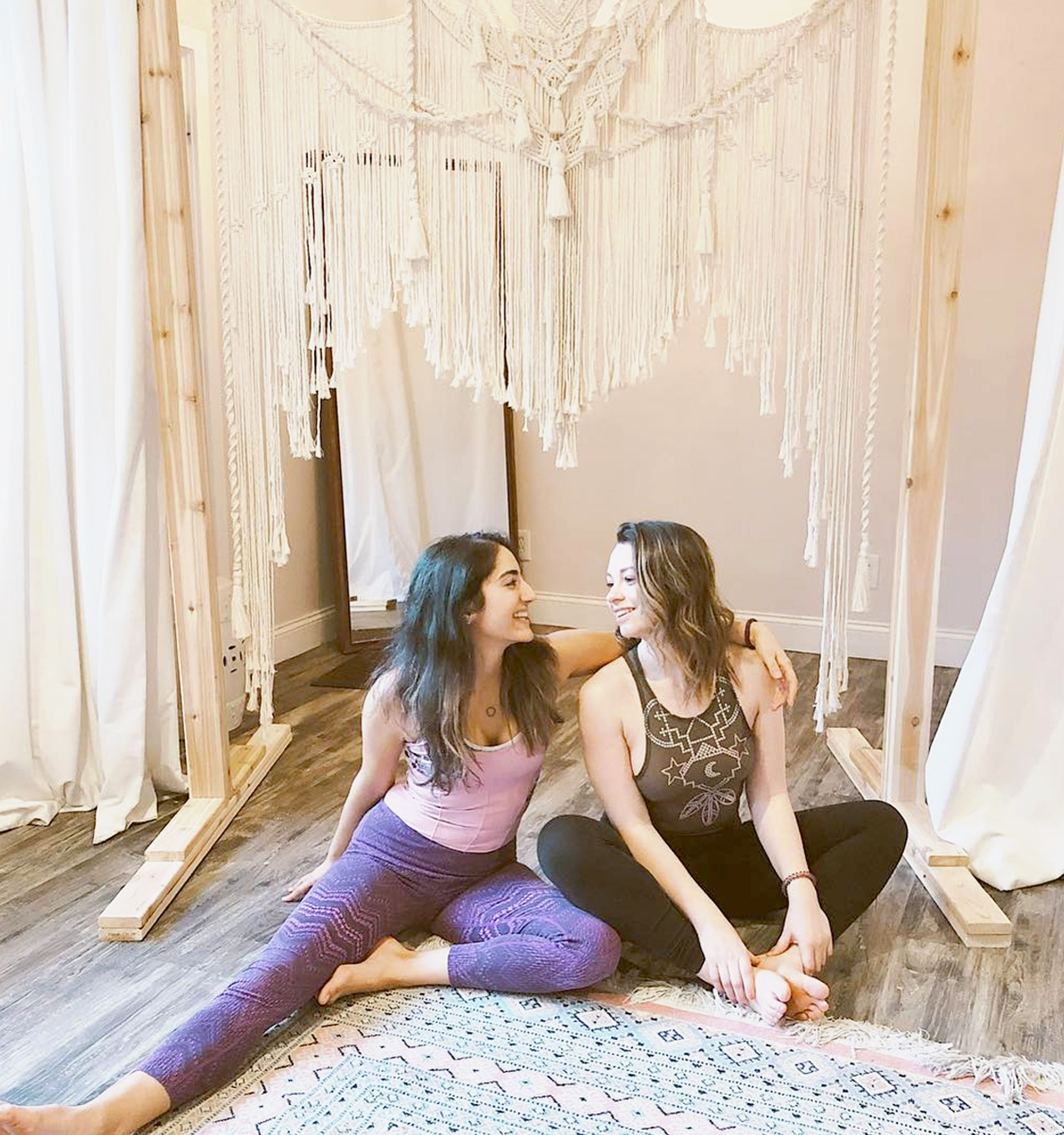 Two women in yoga clothes sitting on a patterned rug in a bright boho wellness studio, smiling under a large macramé wall hanging on wood floors.