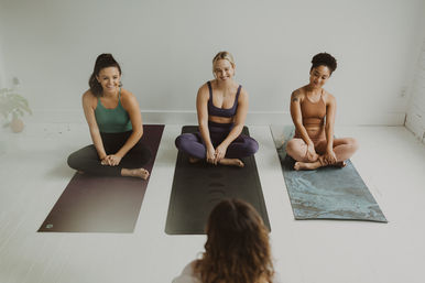 Three women seated cross-legged on yoga mats in a bright, minimalist yoga studio, smiling at their instructor during a group class.