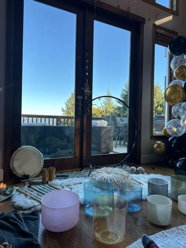 Sound bath setup with colorful crystal singing bowls, drum, cushions and candles on a wooden floor inside, glass patio doors opening to a deck with outdoor seating and evergreen trees under a clear blue sky, black-and-gold balloons at right.
