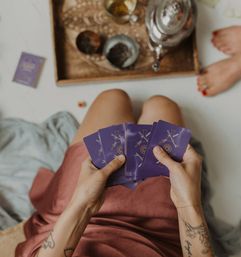 Overhead view of hands fanning purple tarot cards during a cozy at-home tarot reading, wooden tray with teapot and cups nearby, tattooed forearms and painted toes visible