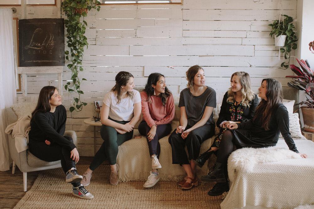 Six women having a relaxed, casual chat in a cozy farmhouse-style living room with white shiplap walls, hanging plants, textured rugs and soft natural light.