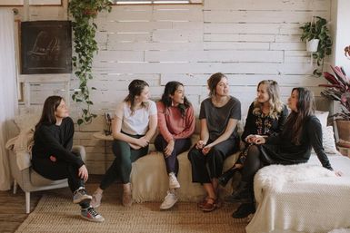 Six women having a relaxed, casual chat in a cozy farmhouse-style living room with white shiplap walls, hanging plants, textured rugs and soft natural light.