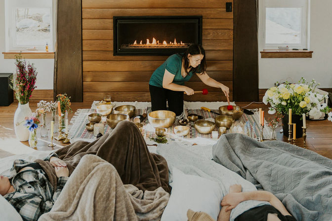 Facilitator playing Tibetan singing bowls during a cozy sound bath session by a fireplace as participants rest under blankets amid candles, flowers, and rugs