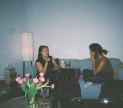 Two people chatting on a blue couch in a cozy living room, drinks on a small table, pink tulips in a vase on the coffee table, floor lamp and stacked books nearby.