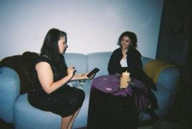 Two women seated on a light-blue sofa in a dim lounge, one jotting notes on a tablet while talking with the other across a small round table draped in purple cloth with a lit candle.