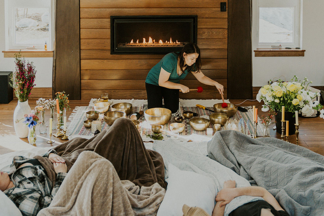 Facilitator leads a cozy living room sound bath by the fireplace, playing singing bowls with mallets while participants relax under blankets amid candles and flowers.