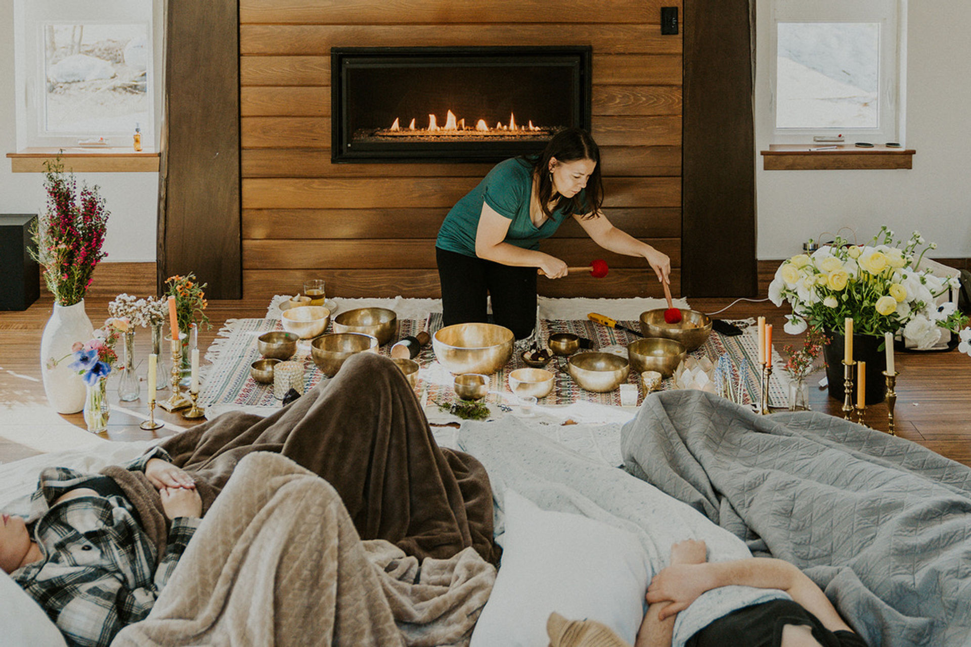 Sound bath session with instructor playing Tibetan singing bowls on a woven rug in a cozy living room by a wood-paneled fireplace, participants resting under blankets for meditation and relaxation.