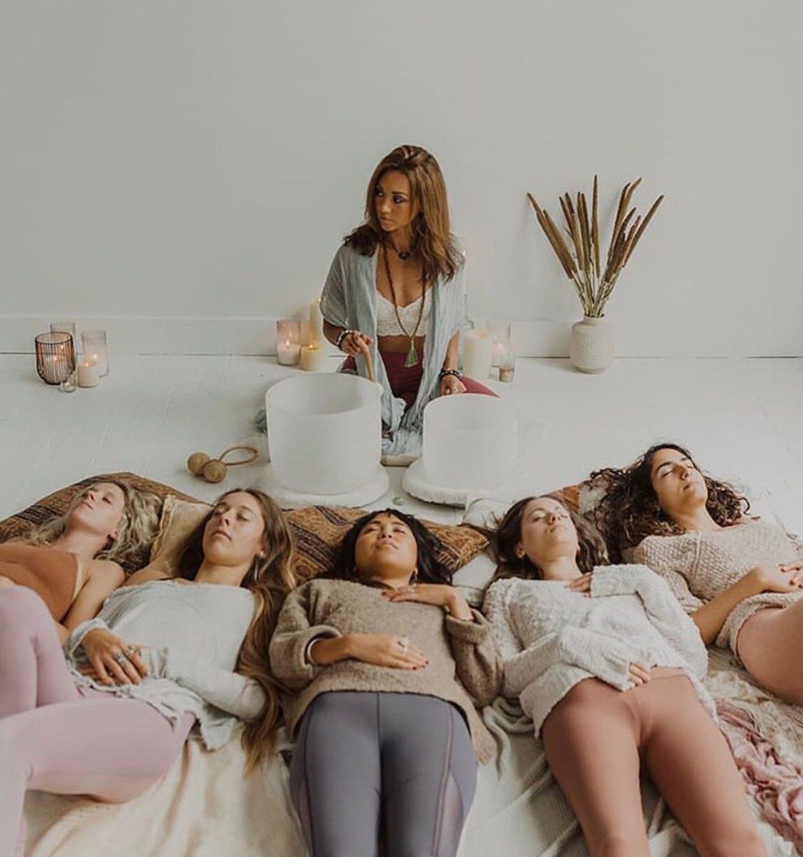 Five women lying with eyes closed in a bright minimalist wellness studio during a guided sound bath meditation, facilitator playing crystal singing bowls surrounded by lit candles and dried plants