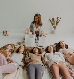 Five women lying with eyes closed in a bright minimalist wellness studio during a guided sound bath meditation, facilitator playing crystal singing bowls surrounded by lit candles and dried plants