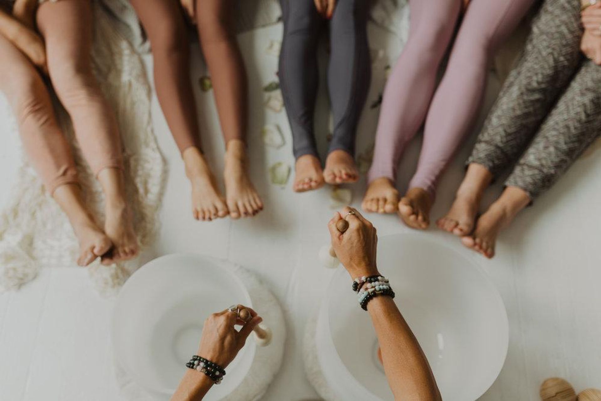 Overhead view of a group sound bath in a bright studio — bare feet and colorful leggings encircling two crystal singing bowls being played with mallets