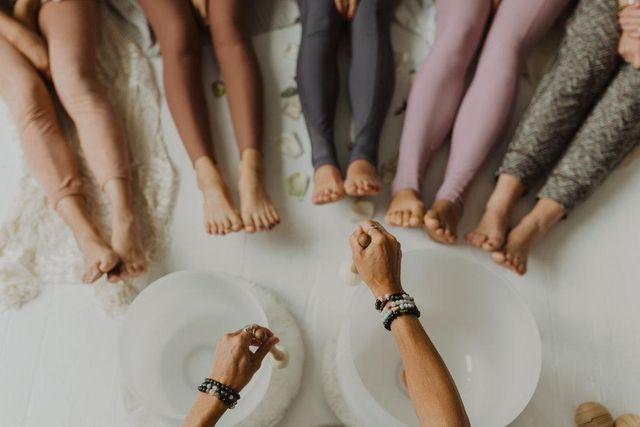 Overhead view of a group sound bath in a bright studio — bare feet and colorful leggings encircling two crystal singing bowls being played with mallets