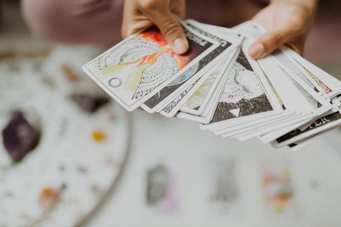 Close-up of hands fanning illustrated tarot cards for a tarot reading over a white table scattered with crystals and spiritual tools
