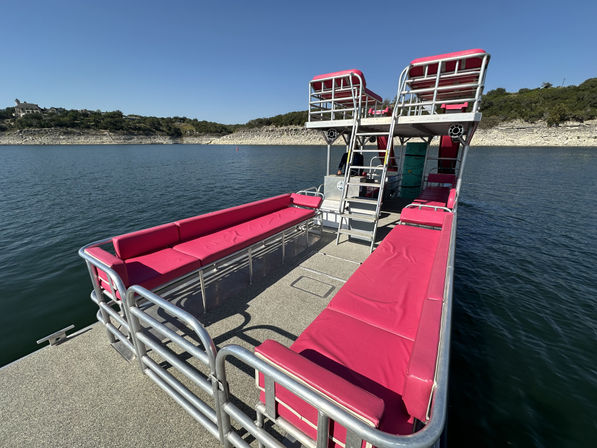 Double-decker pontoon boat with vibrant pink bench cushions and metal railings, floating on a calm lake near a rocky shoreline under a clear blue sky.
