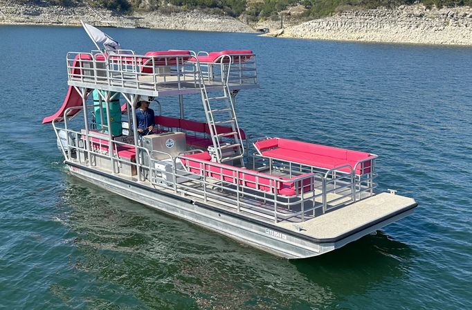 Pontoon party boat with bright pink cushions, upper-deck slide and ladder cruising on a clear blue lake with rocky shoreline