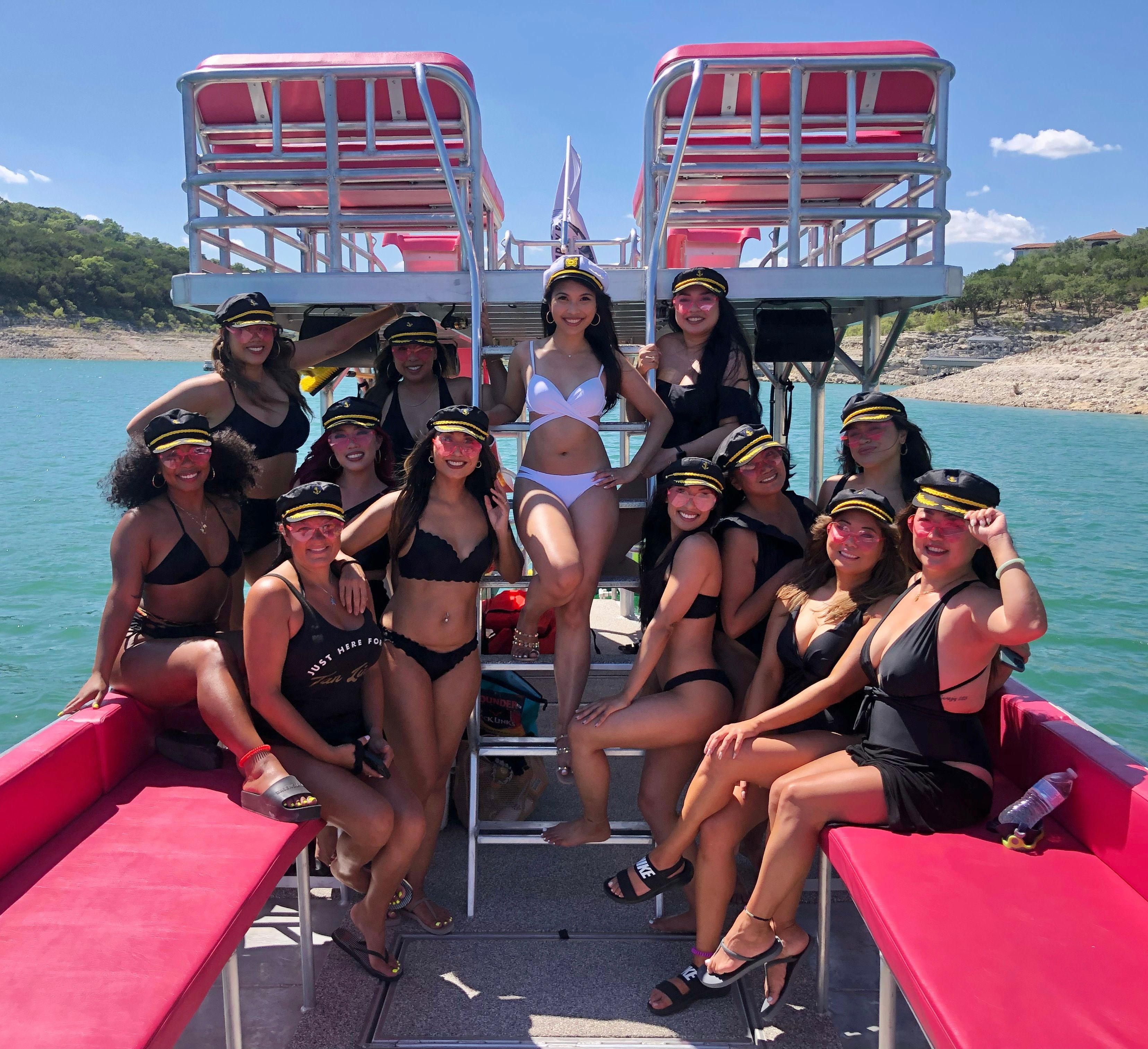 Smiling group of women in black and white swimsuits and captain hats posing on a pink-decked double-decker pontoon boat on a turquoise lake with rocky, tree-lined shoreline and blue sky