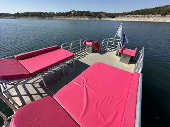 Pontoon boat sundeck with bright pink lounge cushions and metal railings, floating on a calm lake with rocky, tree-lined shoreline under a clear blue sky