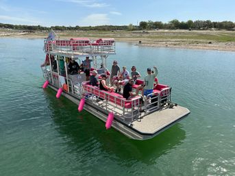 Pontoon boat with bright pink seats and fenders carrying a cheerful group of passengers on a calm lake near a sandy shoreline on a sunny day.