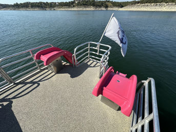 Pink twin water slides on a pontoon-style deck with metal railings and a white nautical flag, overlooking a calm blue lake and rocky tree-lined shoreline.