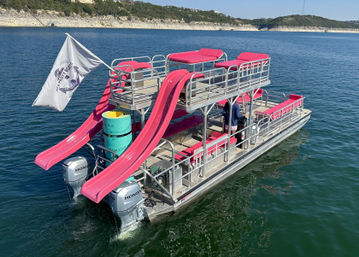 Double-decker pontoon boat with bright pink water slides and matching lounge cushions, twin outboard motors gliding on a clear lake with rocky, tree-covered shoreline under a sunny sky.
