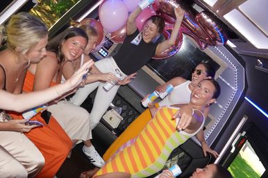 Friends celebrating inside a party bus, laughing and dancing with canned drinks, pink balloons and rose-gold number balloons under colorful LED lighting.