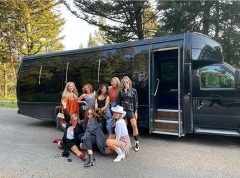 Eight women posing in front of a black party bus parked on a tree-lined road, wearing casual and western-style outfits with boots and a white cowboy hat, smiling for a group photo.