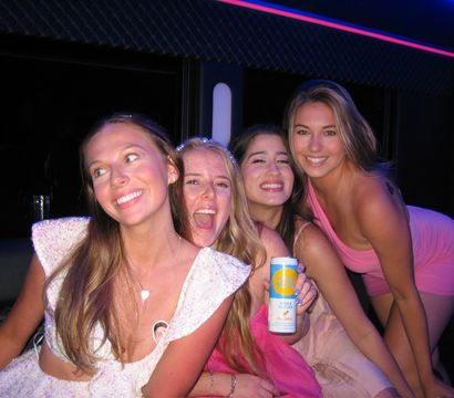Four smiling young women enjoying a neon-lit party bus night out, laughing and posing together while one holds a canned vodka seltzer.