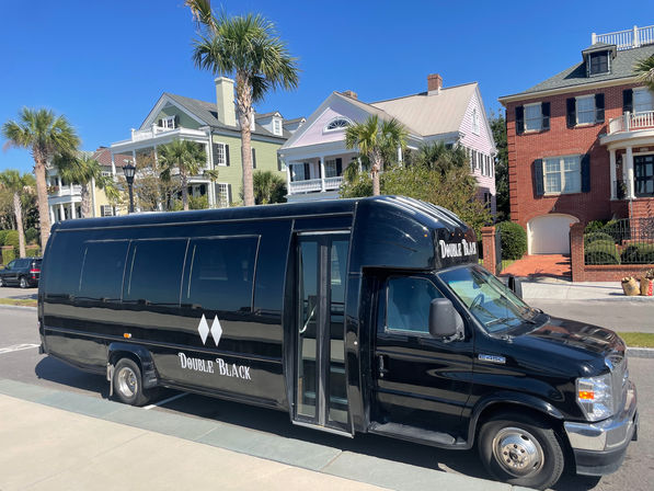Black shuttle bus parked on a palm-lined coastal street in front of pastel-colored Victorian-style houses under a sunny blue sky.