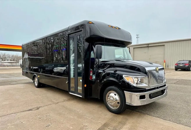 Sleek black commercial shuttle bus with chrome grille and hubcaps parked on a concrete lot in front of a warehouse, glossy side reflecting trees and nearby cars.