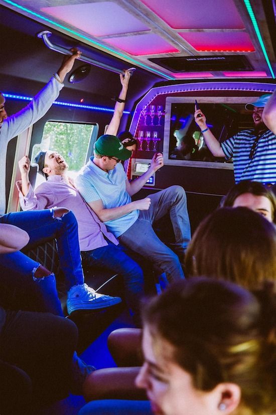 Friends dancing and laughing inside a neon-lit party bus with colorful LED ceiling lights and a small bar area.